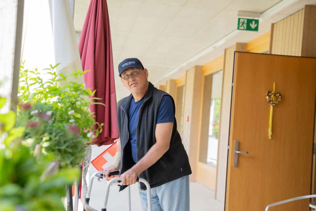 Ein Mann in blauem Hemd, Weste und Mütze steht mit einem Rollator in einem Flur neben einer offenen Tür. Links von ihm stehen Pflanzen und ein roter Regenschirm. Über der Tür ist ein Notausgangsschild zu sehen.