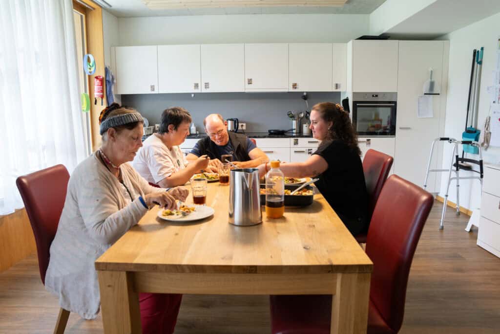 Vier Personen sitzen um einen hölzernen Esstisch in einer modernen Küche. Sie essen gemeinsam, auf dem Tisch stehen Geschirr und Getränke. Die Küche ist mit weißen Schränken und Geräten ausgestattet und der Raum hat große Fenster, die für Tageslicht sorgen.