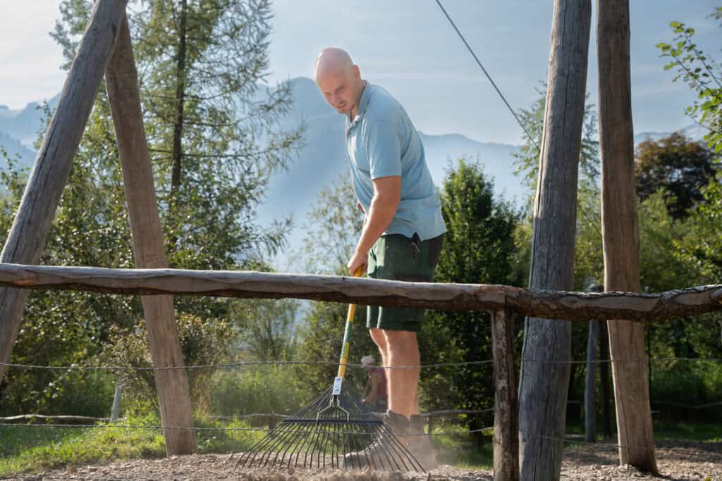 Eine Person harkt Kies in einem umzäunten Bereich mit Holzpfosten für Gleichgewichtsübungen, umgeben von Bäumen und Bergen im Hintergrund an einem sonnigen Tag.